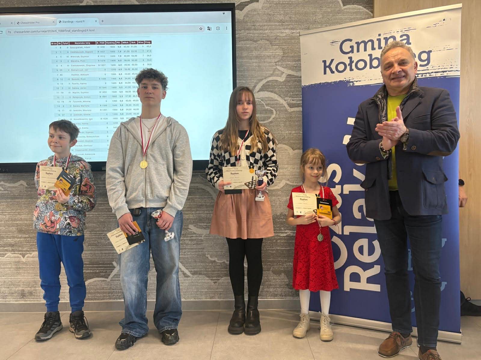 Dzieci stoją z medalami i dyplomami podczas wręczenia nagr&oacute;d w turnieju szachowym; obok dorosły bije brawo. W tle ekran z wynikami oraz baner &bdquo;Gmina Kołobrzeg&rdquo;, scena przedstawia podsumowanie i nagrodzenie uczestnik&oacute;w.